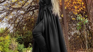 Woman in a floor-length black wrap coat with a tied waist sash and black boots standing among golden autumn leaves