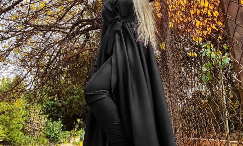 Woman in a floor-length black wrap coat with a tied waist sash and black boots standing among golden autumn leaves