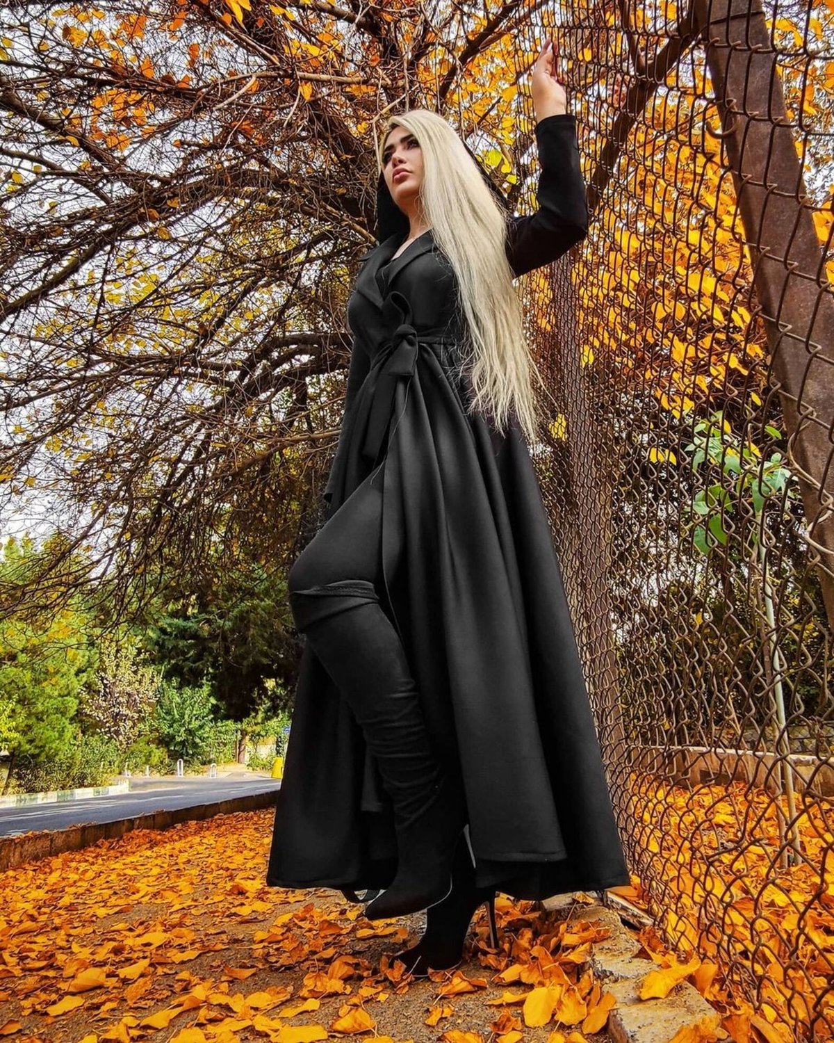 Woman in a floor-length black wrap coat with a tied waist sash and black boots standing among golden autumn leaves