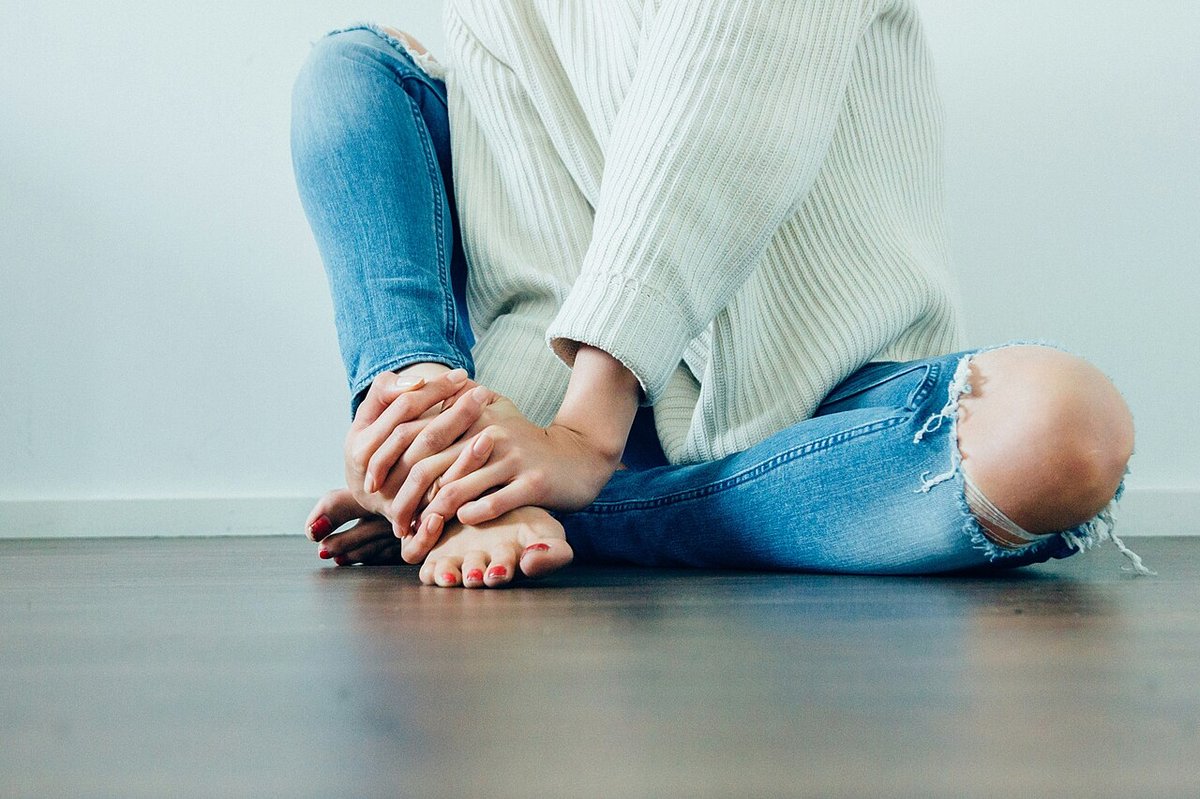 Woman seated cross-legged on a wooden floor wearing a cream chunky knit sweater and ripped blue jeans with a torn knee, barefoot