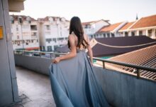Back view of a woman in a floor-length grey-blue satin gown with an embellished waistband, holding the skirt on a rooftop terrace