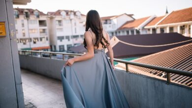 Back view of a woman in a floor-length grey-blue satin gown with an embellished waistband, holding the skirt on a rooftop terrace