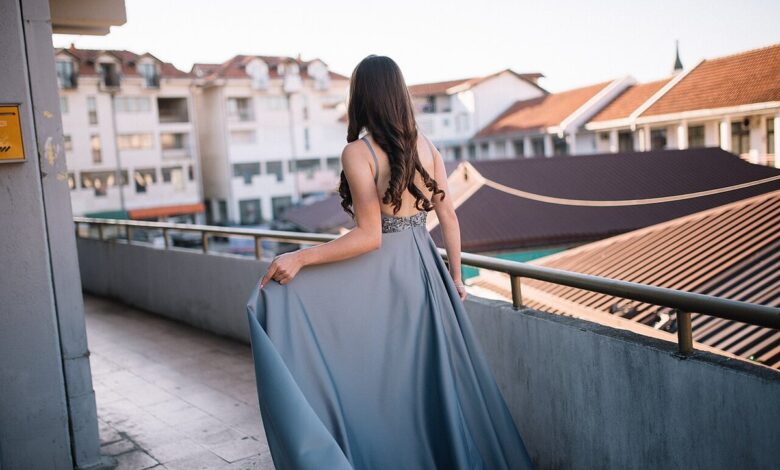 Back view of a woman in a floor-length grey-blue satin gown with an embellished waistband, holding the skirt on a rooftop terrace