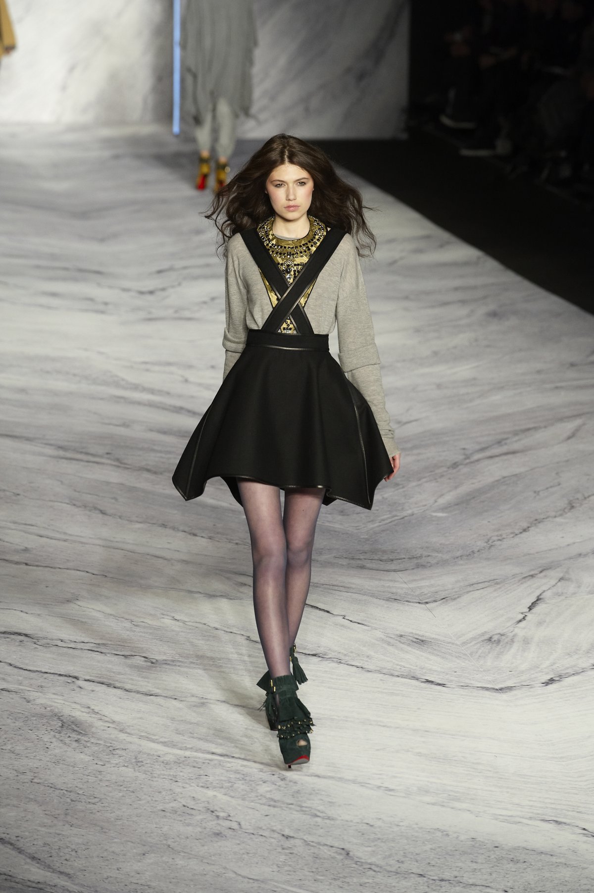 Model walking a runway in a grey knit sweater, black skater skirt with crossed straps, sheer black tights and black fringed ankle boots.