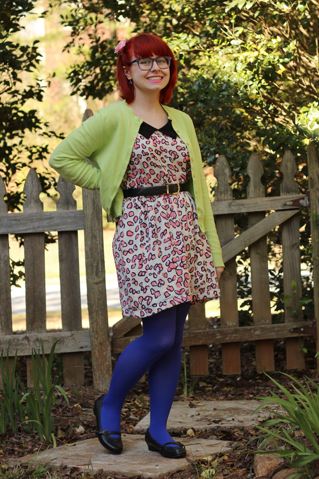 A woman with red hair and glasses wearing a pink leopard print dress, a lime green cardigan, blue tights and black Mary Jane shoes, standing outdoors by a wooden fence.