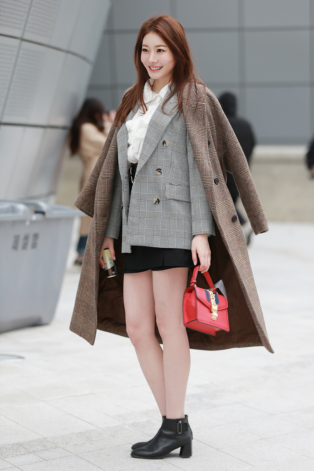 A woman with long auburn hair standing outdoors in a city plaza, wearing a long chocolate-brown plaid wool coat over a grey plaid blazer, white ruffled blouse, black mini skirt and black ankle boots, carrying a small red handbag