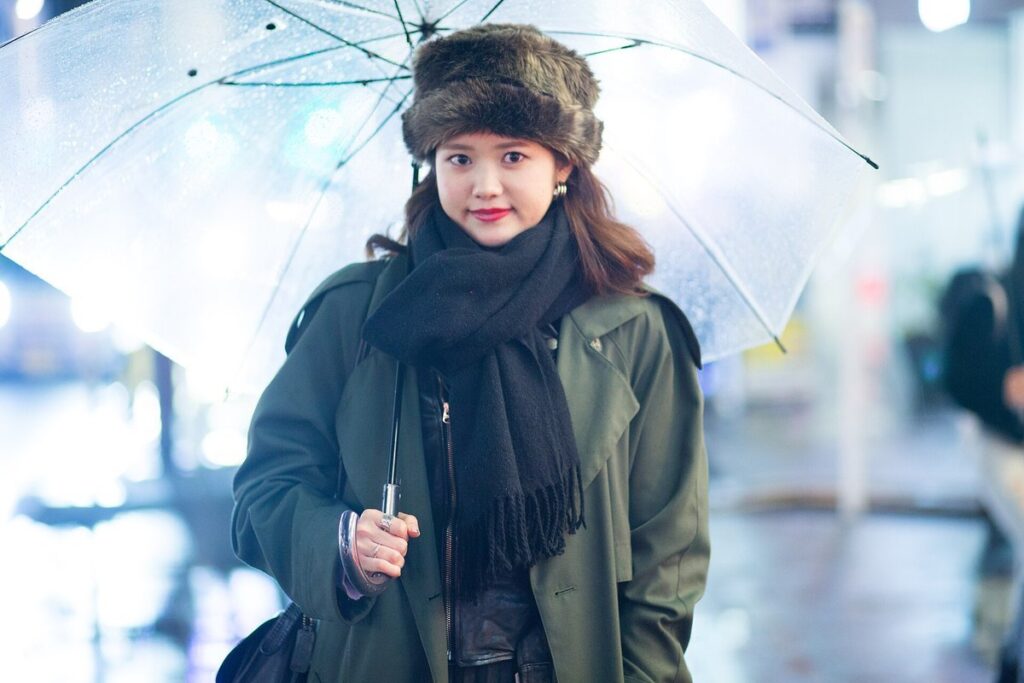 Woman in a fur hat and black scarf wearing an olive green trench coat, holding a clear umbrella on a rainy Harajuku street at night