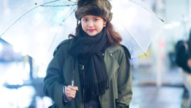 Woman in a fur hat and black scarf wearing an olive green trench coat, holding a clear umbrella on a rainy Harajuku street at night