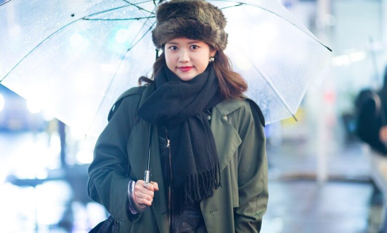 Woman in a fur hat and black scarf wearing an olive green trench coat, holding a clear umbrella on a rainy Harajuku street at night