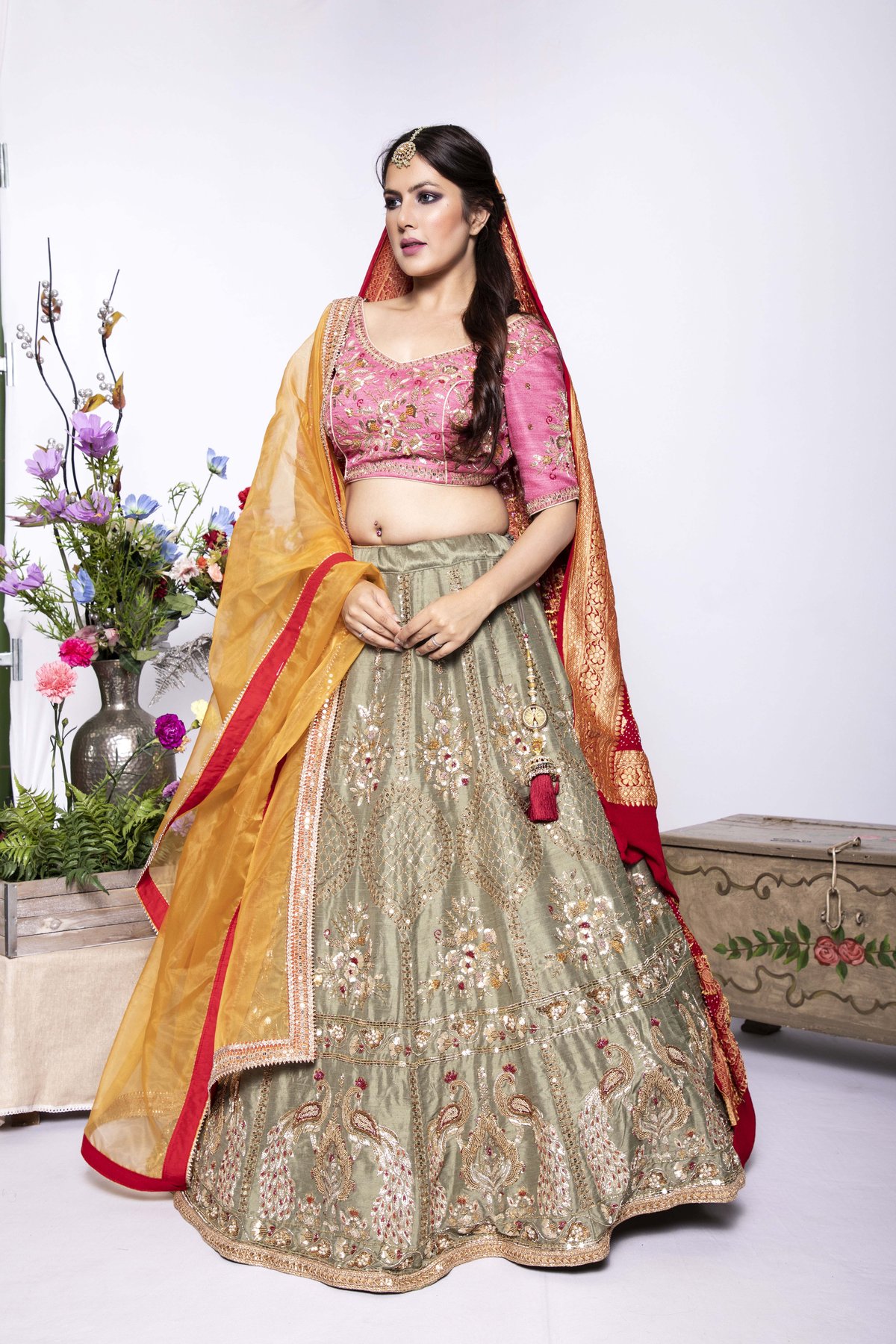 Woman in a blush-pink embroidered choli and olive-sage embroidered flared lehenga with an orange-red dupatta