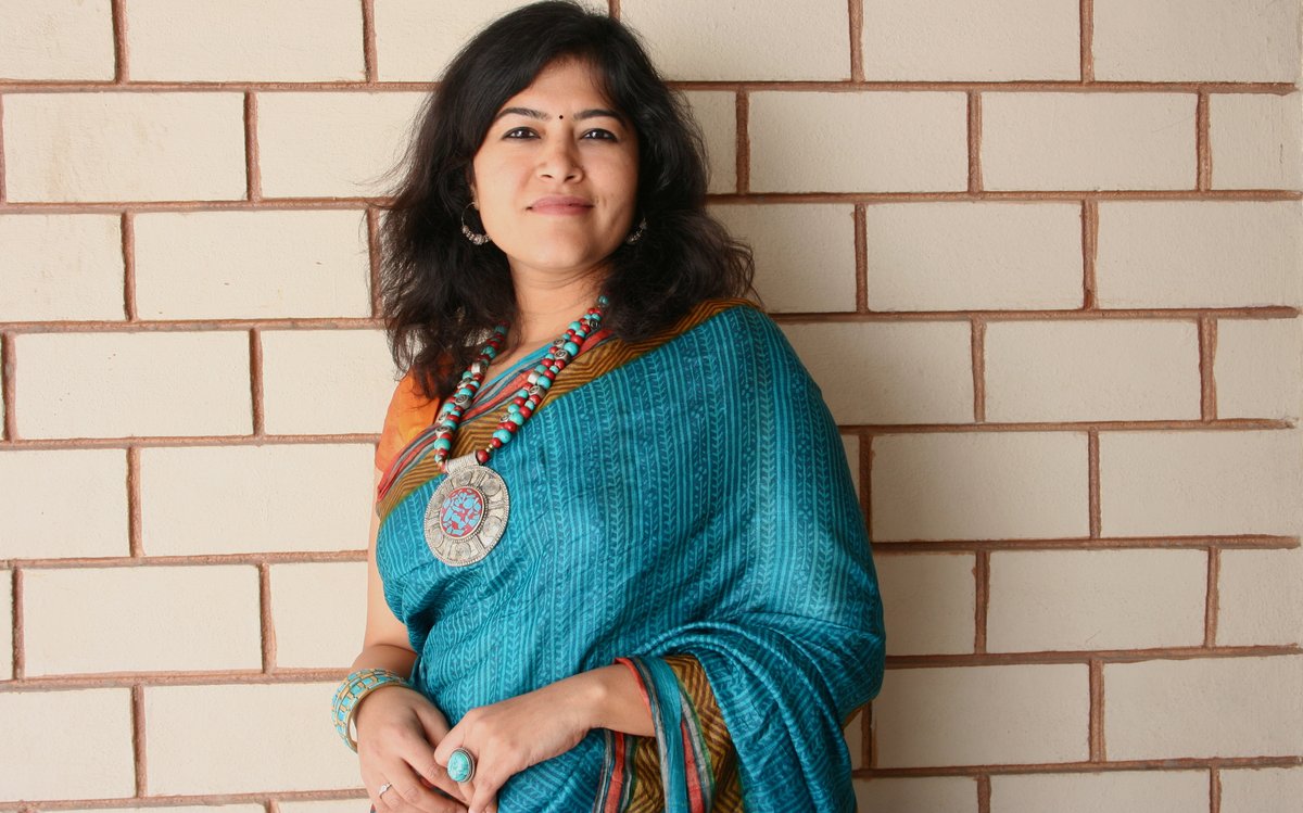 Woman wearing a teal-blue saree with a statement beaded necklace and bangles, standing against a brick wall.