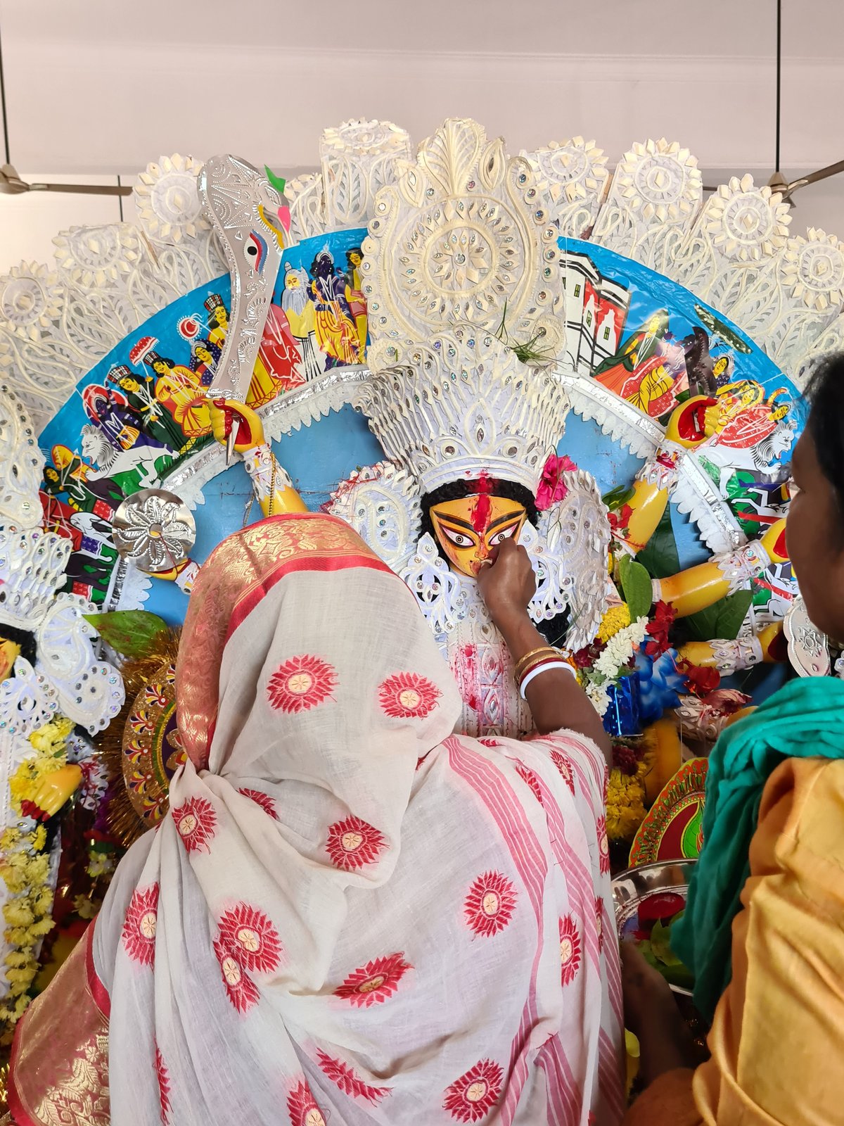 Woman in a white saree with red floral motifs and a gold border leaning over a Durga idol to perform the boron farewell ritual