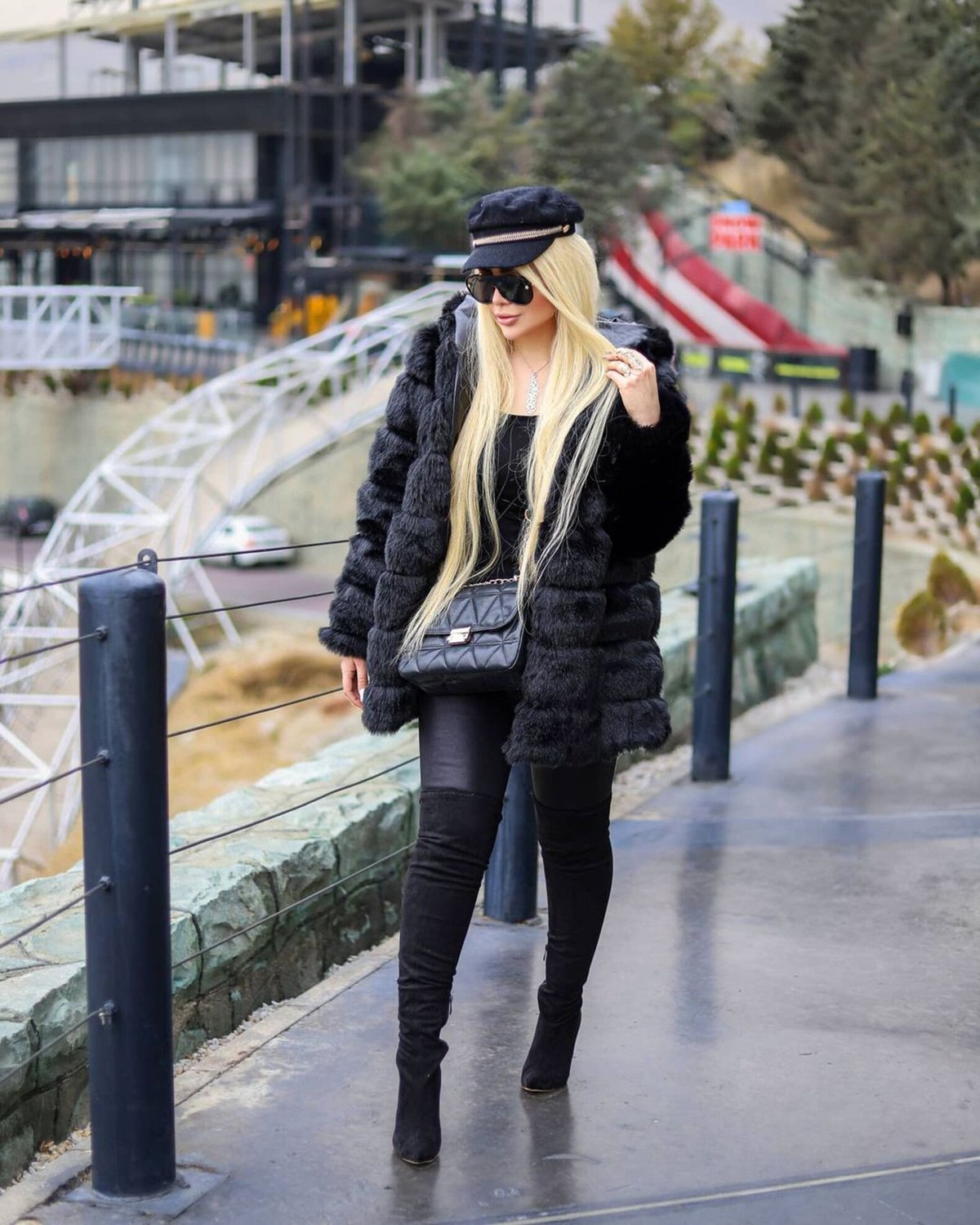 Woman in an all-black winter outfit with a chunky faux-fur coat, over-the-knee boots, flat cap and crossbody bag on a waterfront promenade