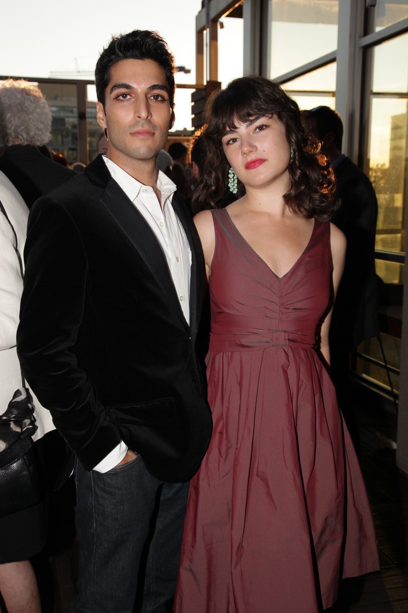 Woman at an indoor evening reception wearing a burgundy sleeveless V-neck fit-and-flare dress with a gathered bodice and a full skirt past the knee, standing beside a man in a black velvet blazer