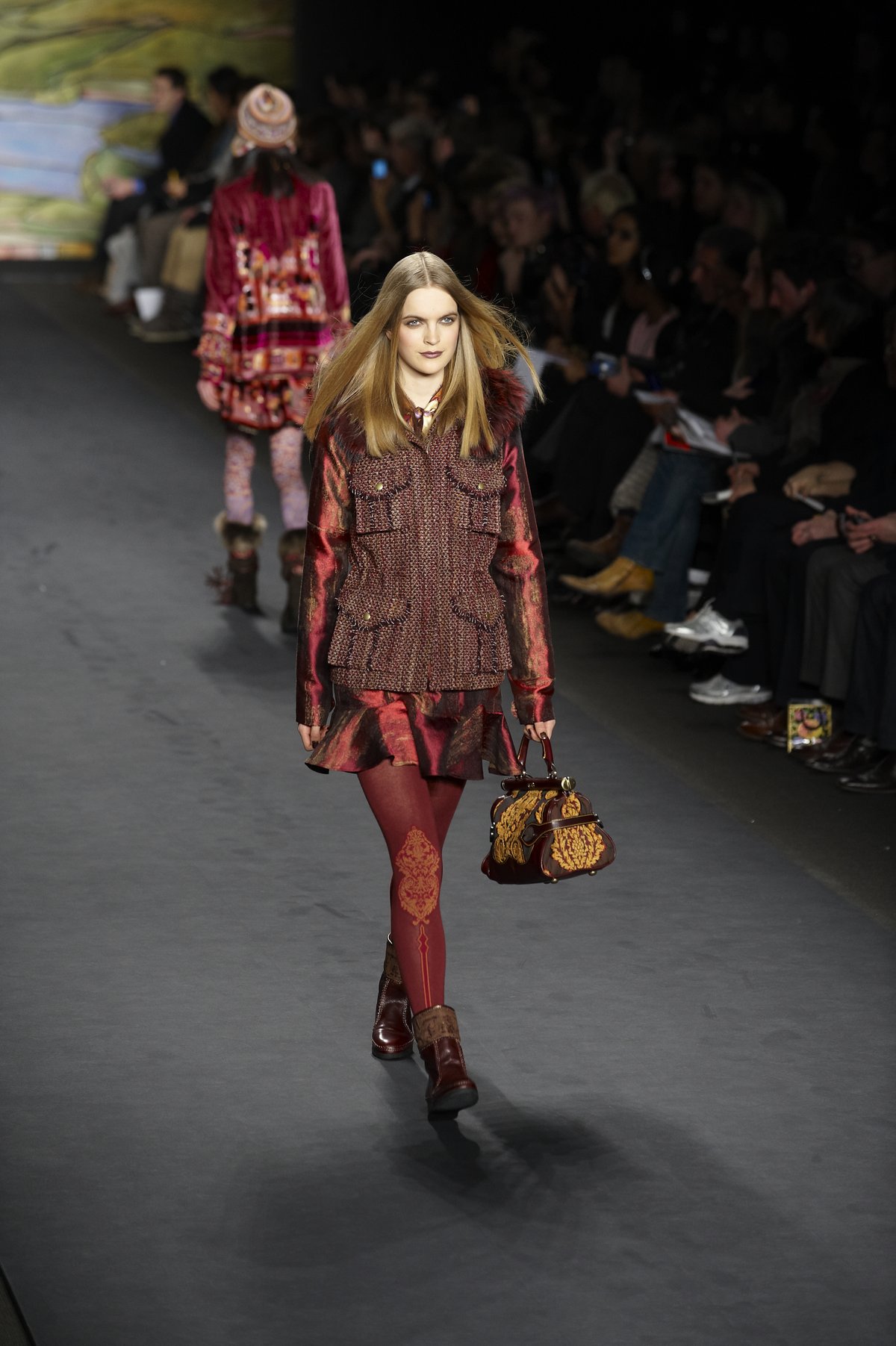 Model on a runway wearing a burgundy tweed jacket with a fur collar, burgundy patterned tights, brown leather boots and a knit beanie, with another model in a beanie behind her.