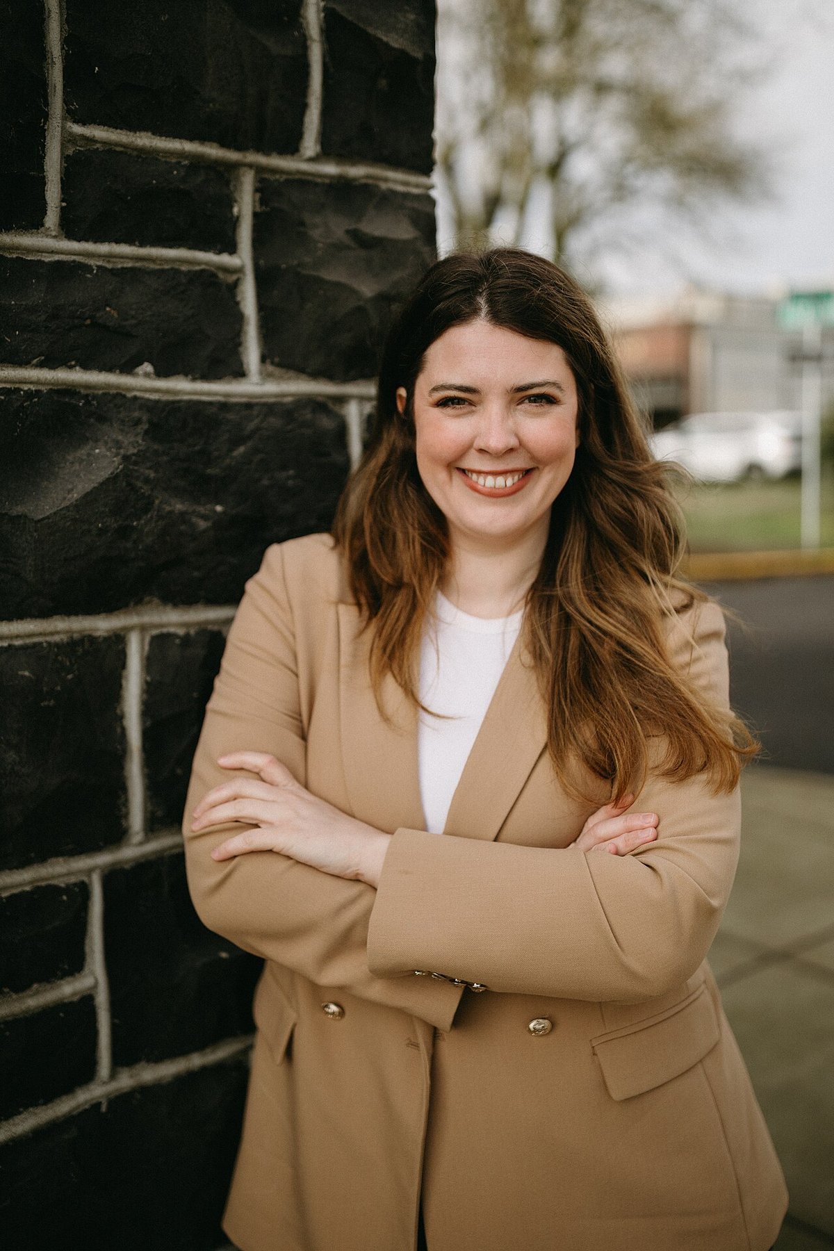 Smiling woman in a double-breasted camel blazer and white top standing by a stone wall
