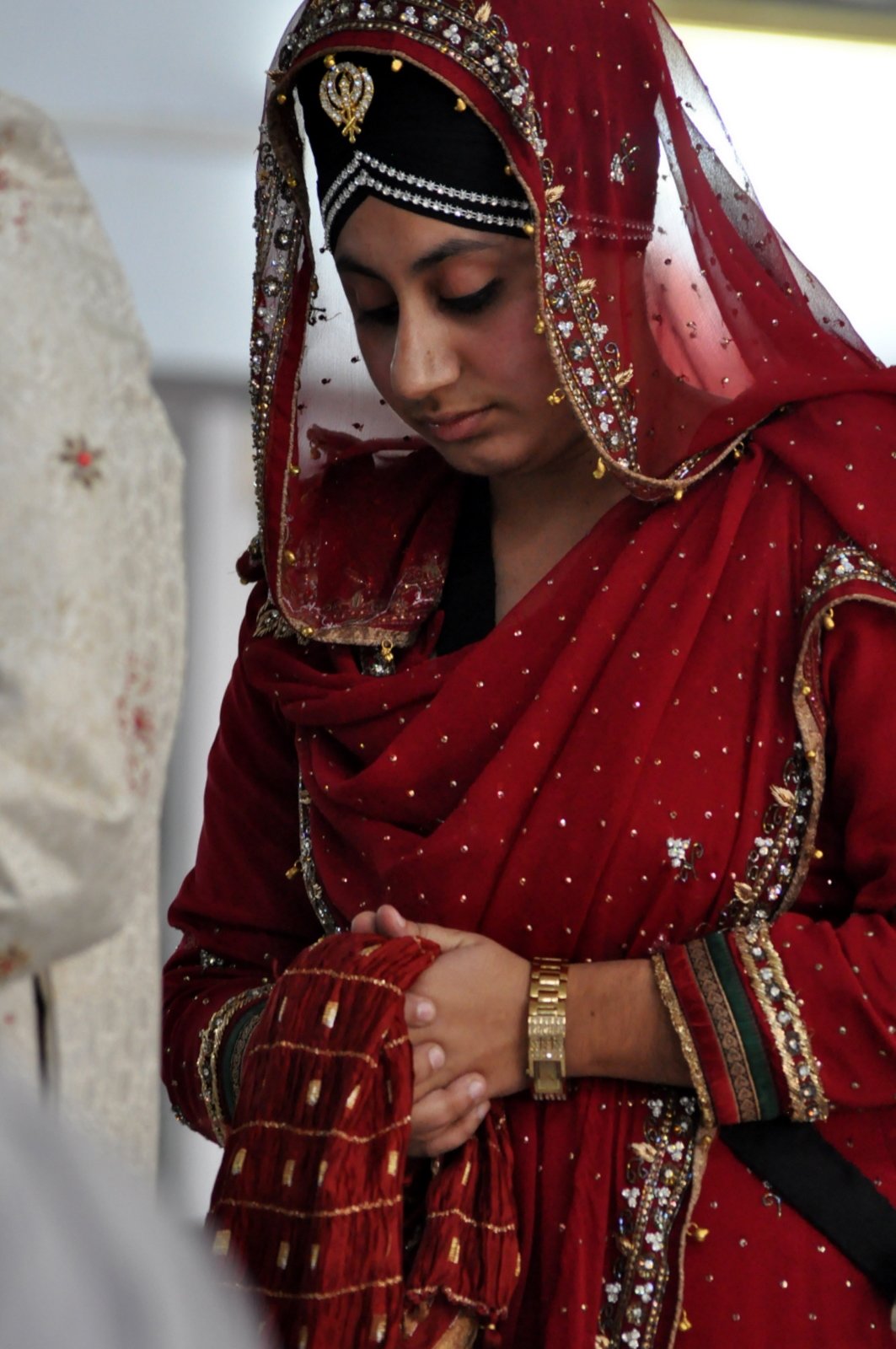 Woman in a deep red embroidered bridal suit with a sheer red dupatta draped over her head, holding a folded red cloth