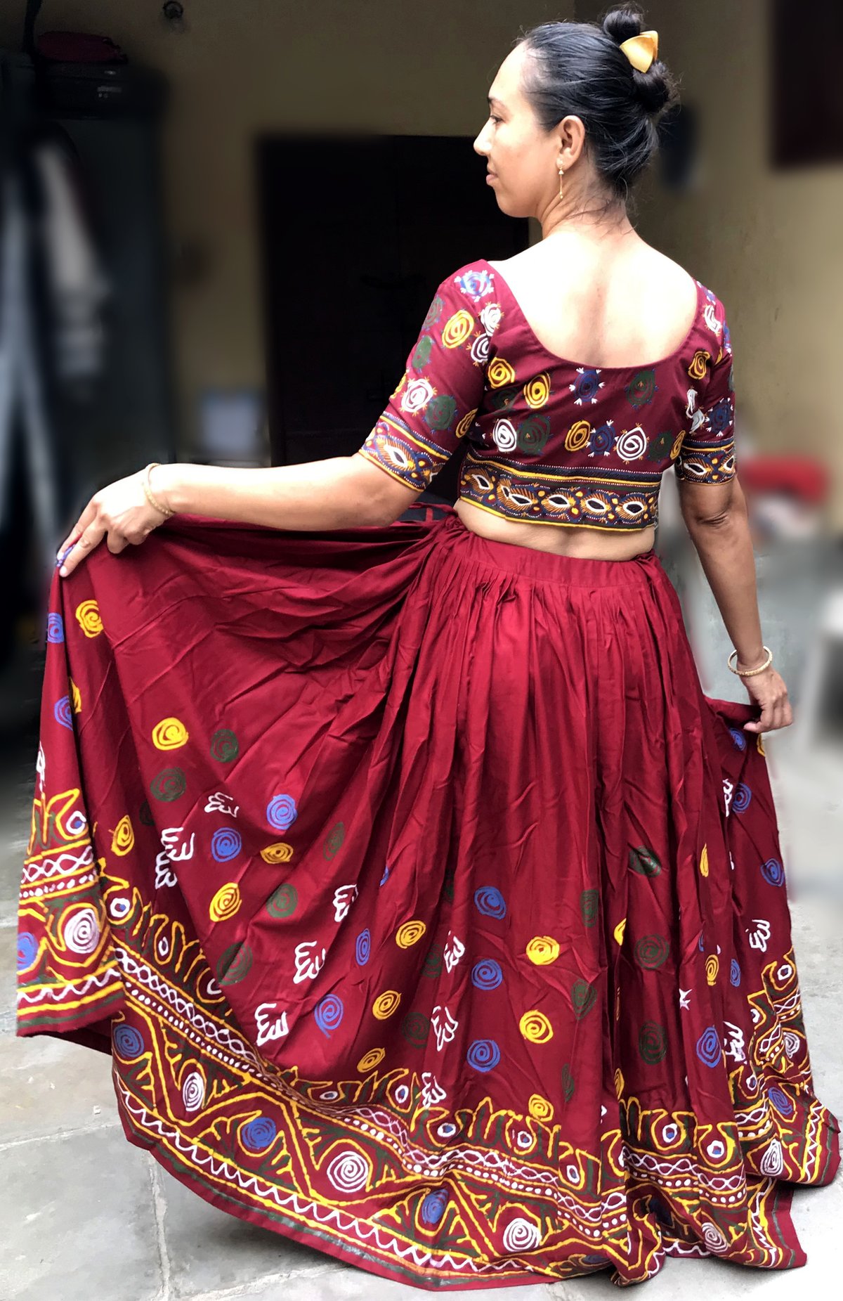 Back view of a woman in a maroon lehenga with colorful Rogan-painting motifs holding out the flared skirt