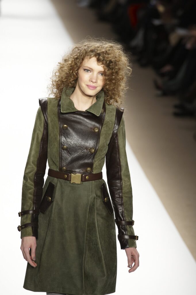 Runway model wearing an olive green suede coatdress with dark brown leather panels and brass buttons