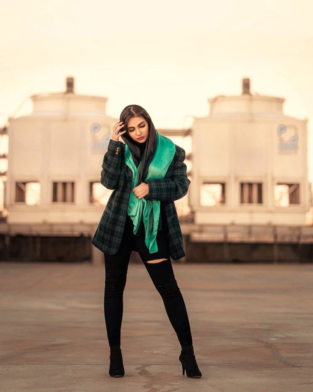 Woman in a dark plaid coat, ripped black skinny jeans and heeled ankle boots, with a black headscarf and emerald green scarf