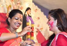 Two smiling women in red sarees applying sindoor to each other during Bijoya Dashami celebrations with a Durga idol behind them