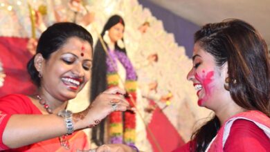 Two smiling women in red sarees applying sindoor to each other during Bijoya Dashami celebrations with a Durga idol behind them