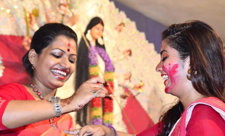 Two smiling women in red sarees applying sindoor to each other during Bijoya Dashami celebrations with a Durga idol behind them