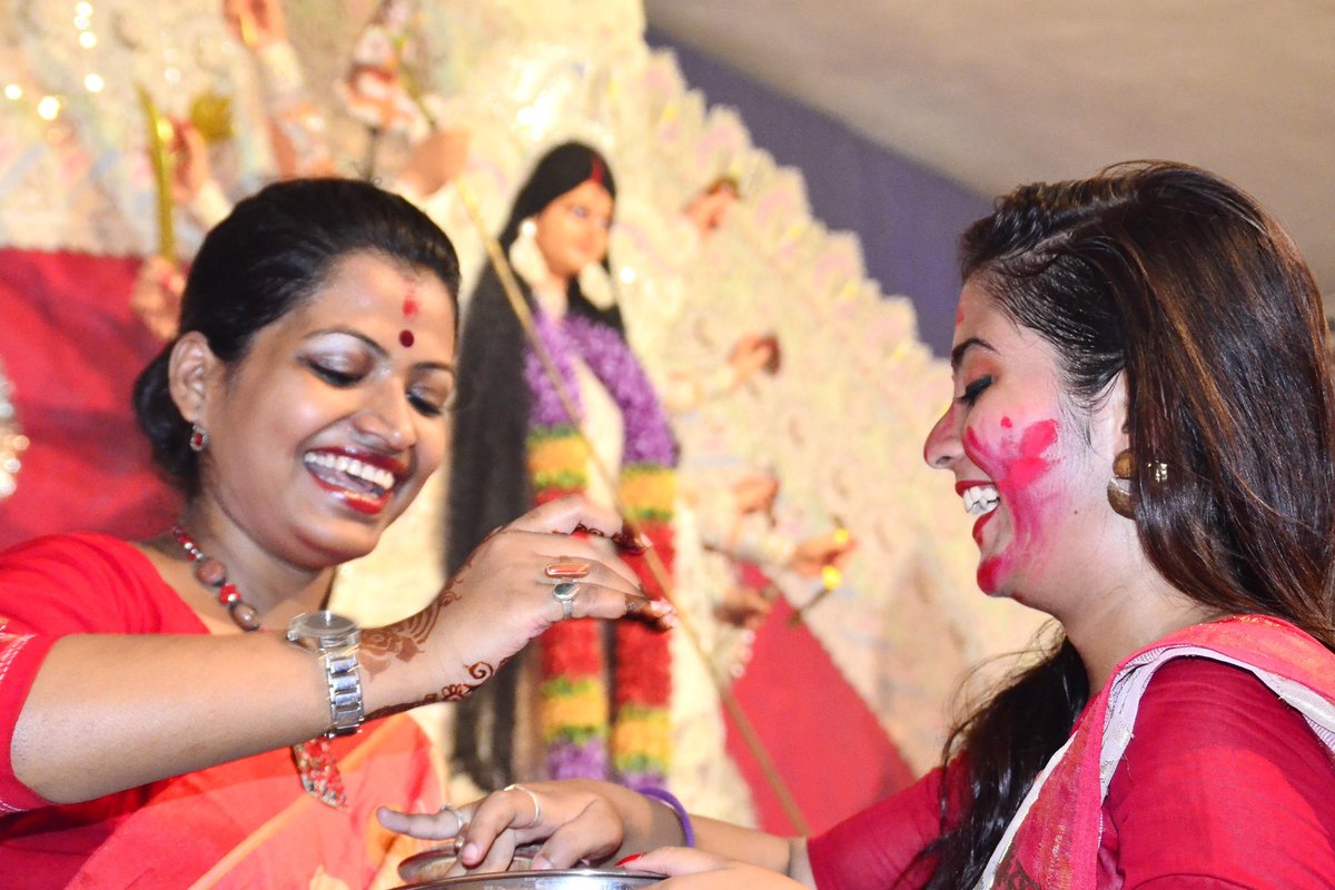 Two smiling women in red sarees applying sindoor to each other during Bijoya Dashami celebrations with a Durga idol behind them