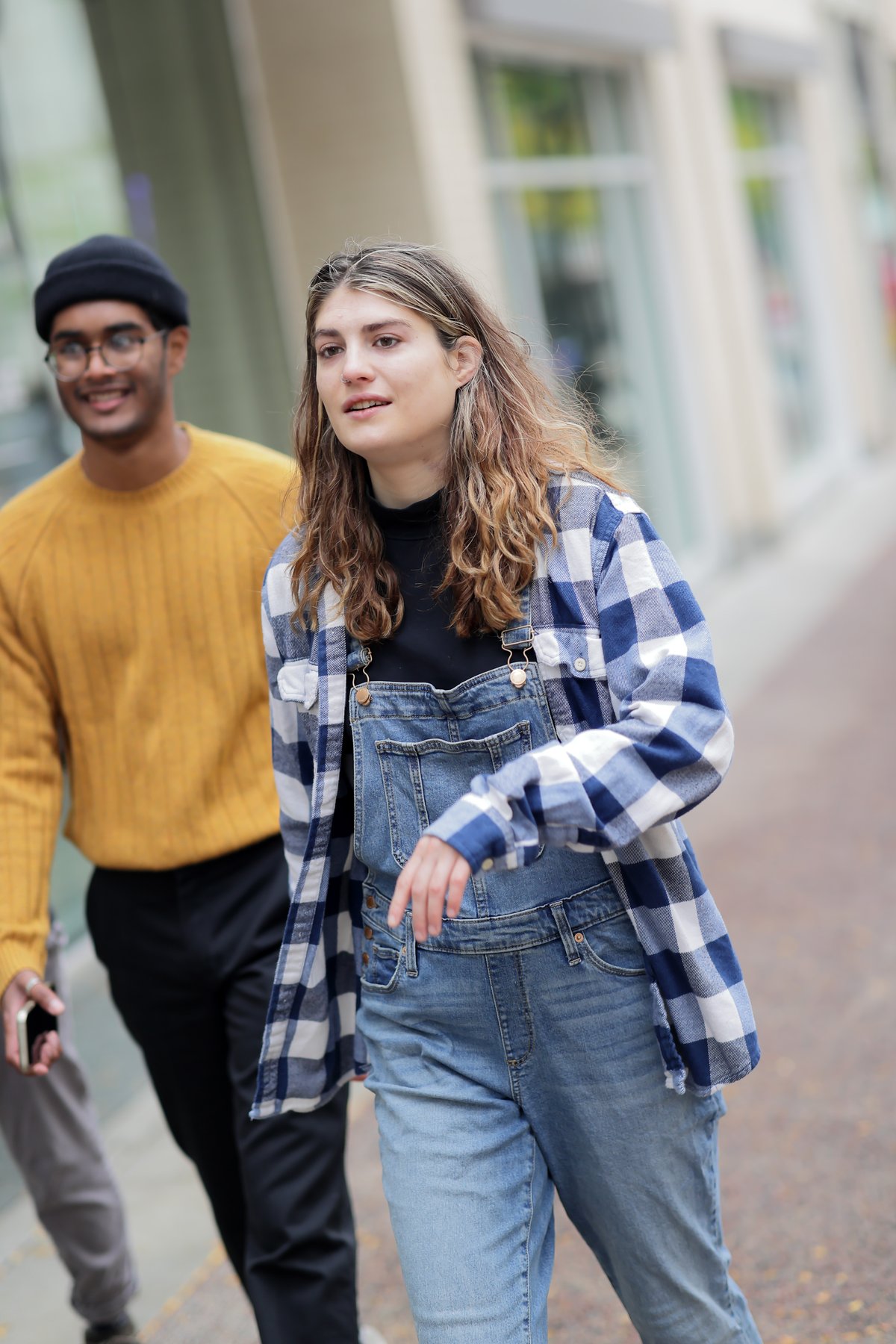 Young woman on a city street wearing a blue and white buffalo plaid flannel shirt open over a black top and denim overalls