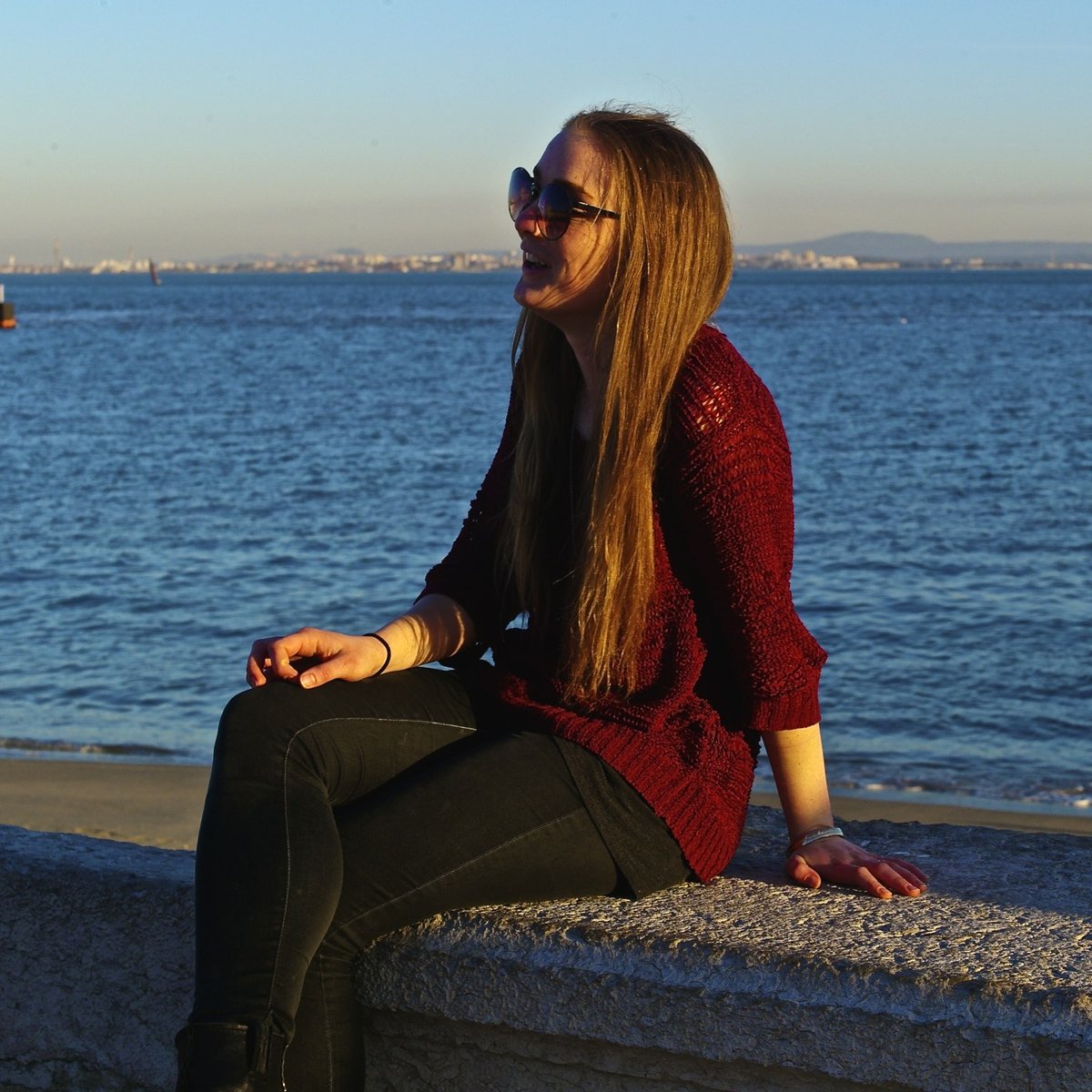Young woman with long brown hair and sunglasses sitting on a stone ledge by the sea at golden hour, wearing a true-burgundy chunky-knit crew-neck sweater with sleeves rolled to three-quarter length and black skinny pants