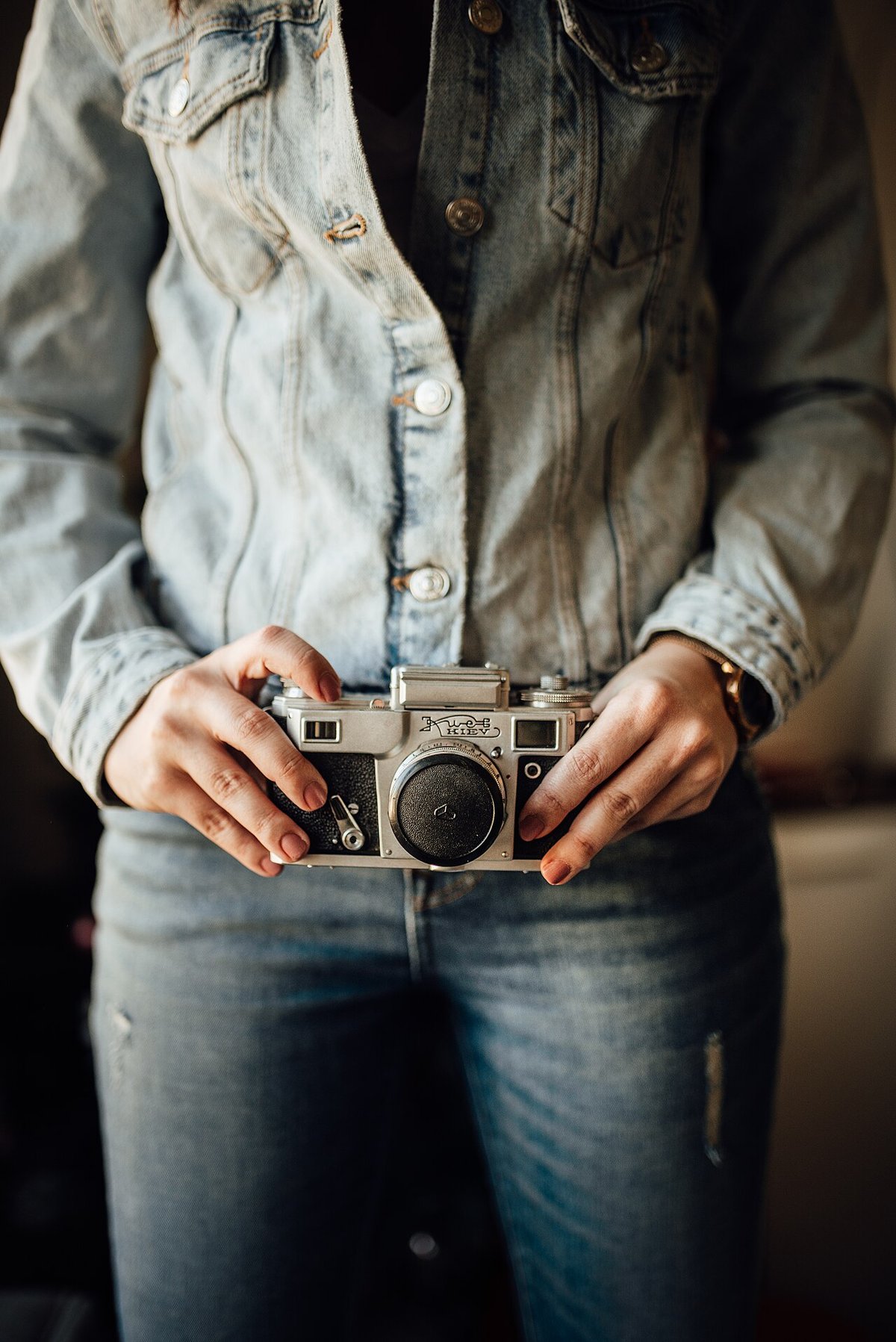 Close crop of a woman in a light-wash denim jacket and medium-blue jeans holding a vintage film camera at her waist
