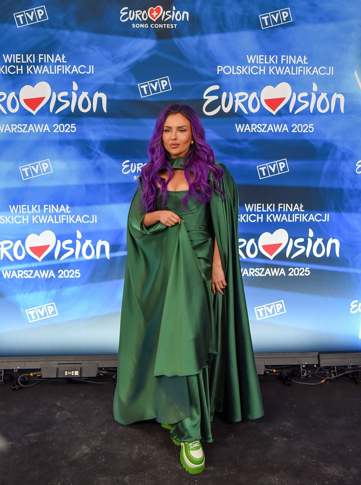 Woman with purple hair wearing a floor-length emerald green satin gown with flowing cape sleeves, standing against a blue Eurovision backdrop