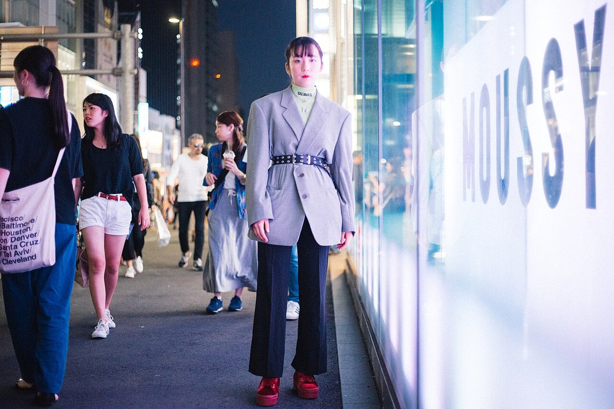 Woman on a city street at night wearing an oversized light grey blazer cinched with a wide black belt, black trousers and red shoes