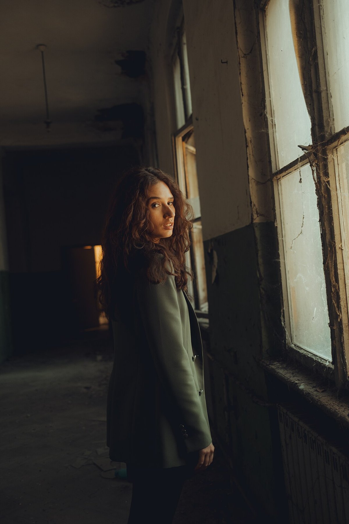 Woman with curly hair in a dark olive blazer standing by a dusty window in an old building