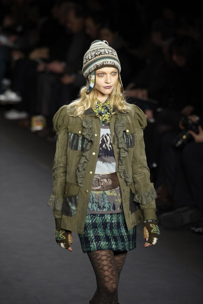 Runway model wearing an olive utility jacket with lace ruffle trim, a knit hat, and a patterned skirt