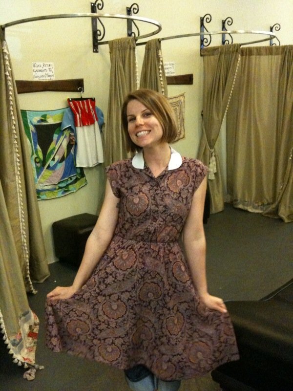 Smiling woman wearing a purple paisley-print dress with a white collar in a dressing room.