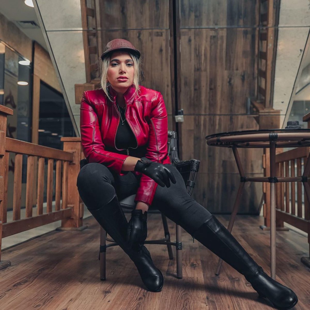 Woman seated indoors wearing a red leather jacket, black leggings, black over-the-knee boots, black gloves and a brown cap