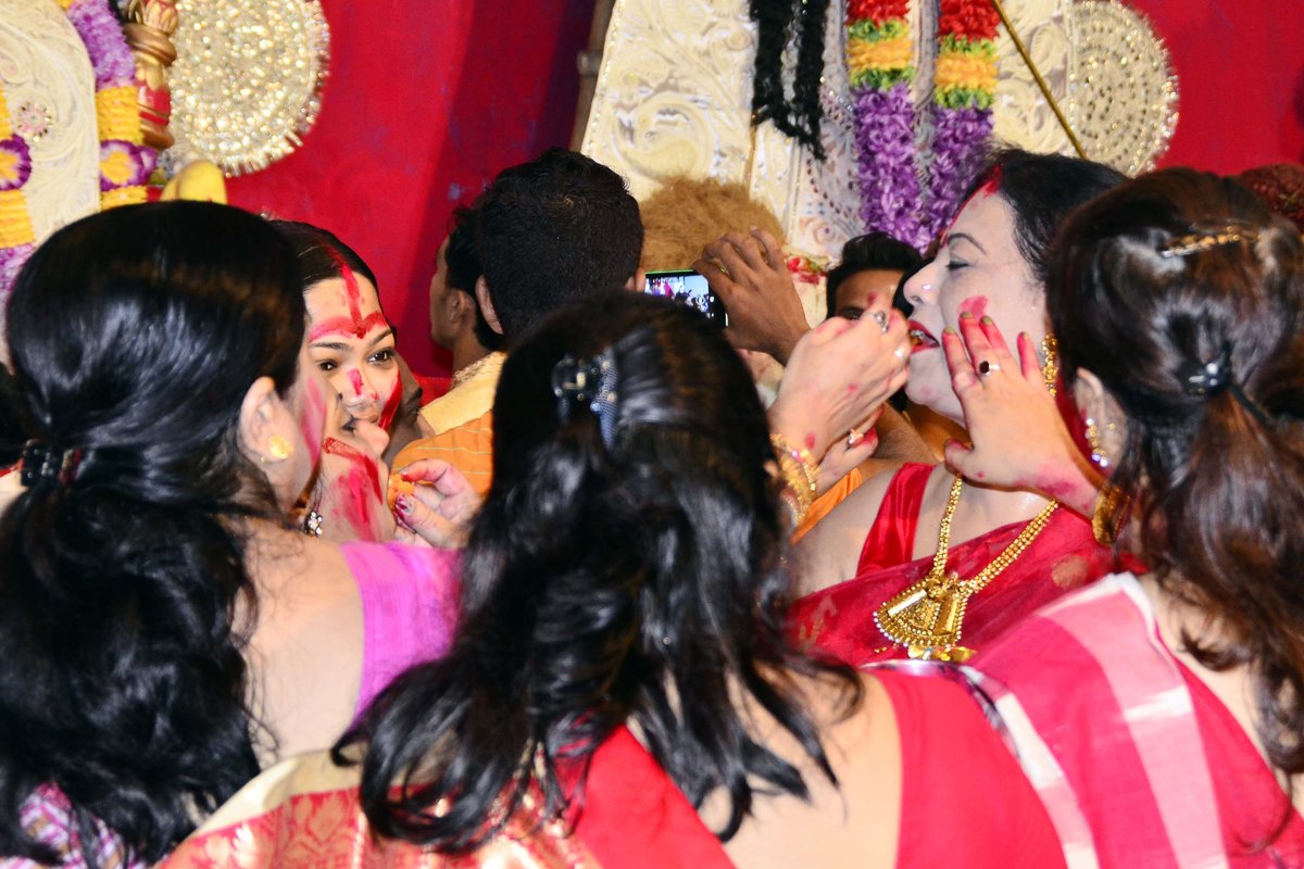 Woman in a red saree wearing a heavy gold necklace having sindoor applied to her face by other women at a Durga Puja pandal