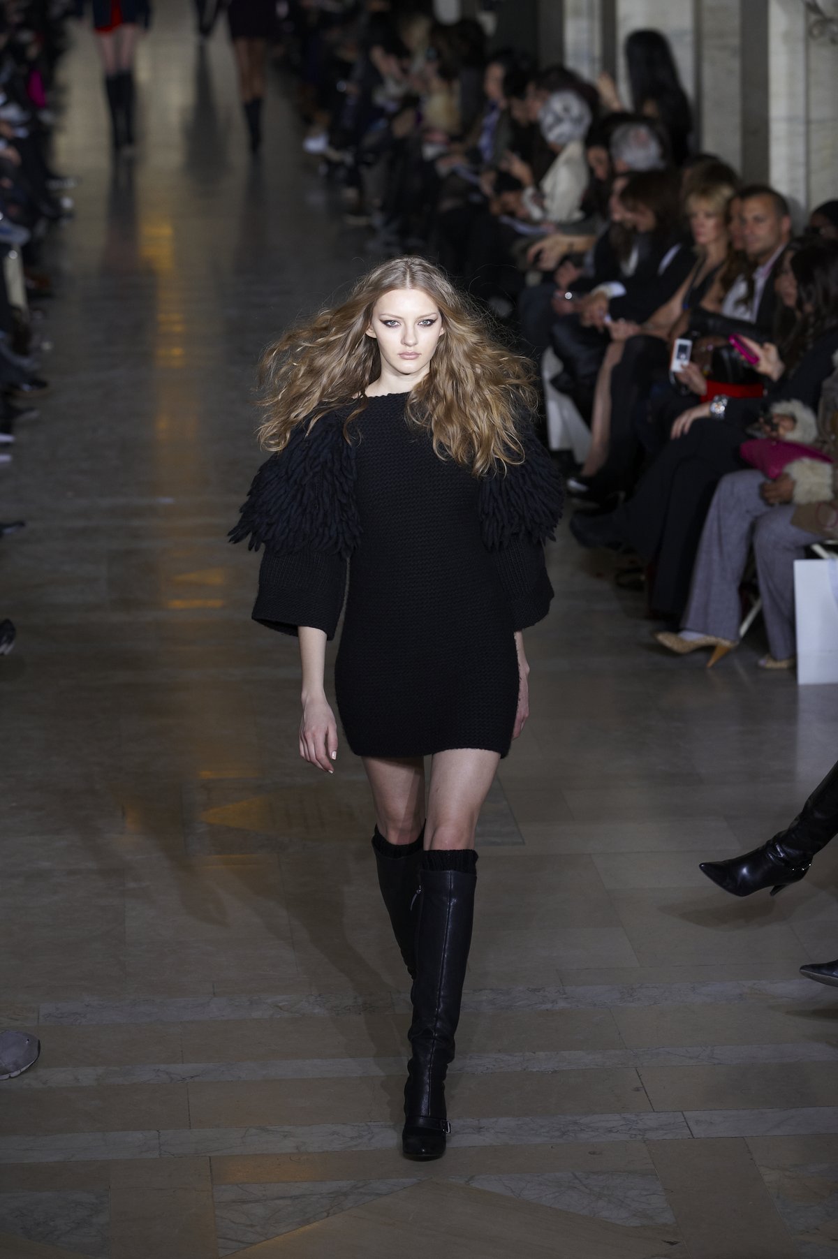 Model on a runway wearing a black knit sweater dress with fringed shoulders and black knee-high boots.
