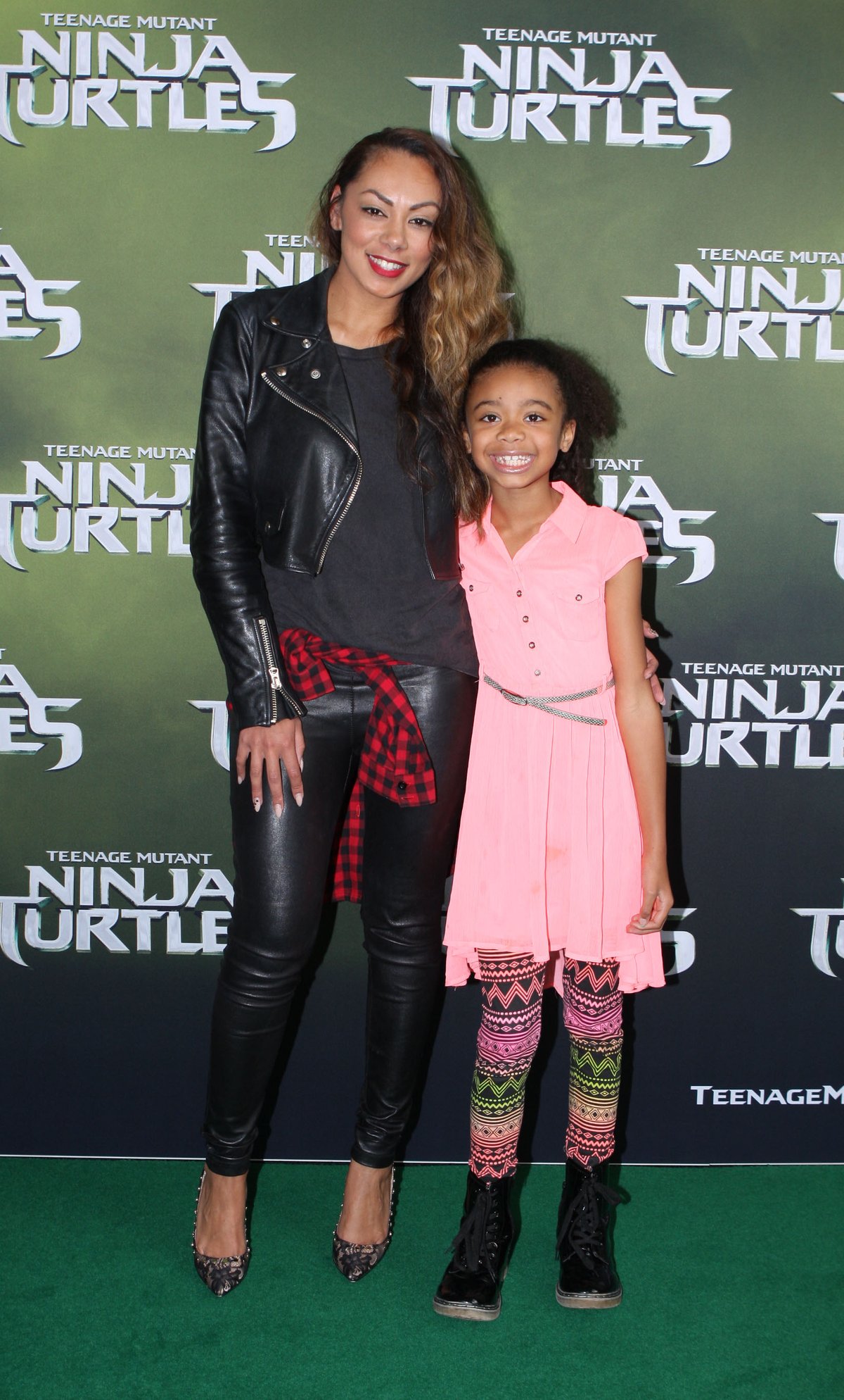 Woman on a premiere red carpet in a black leather biker jacket, black top, tight black leather pants, a red plaid shirt tied at the waist and glittery heels