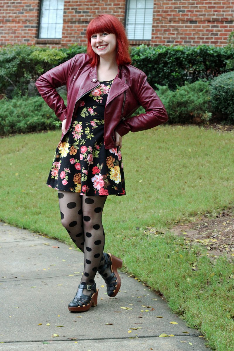 Woman with shoulder-length red hair standing on a sidewalk, wearing an open deep-burgundy faux-leather moto jacket with a diagonal zip over a black fit-and-flare dress printed with large roses, sheer black polka-dot tights, and black strappy platform sandals