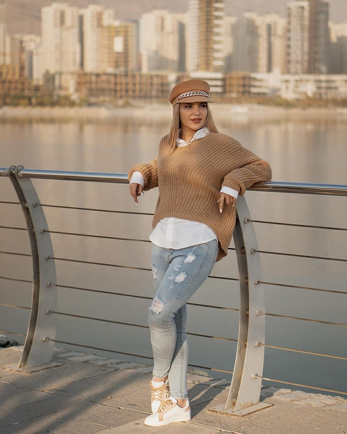 Woman by a lake in an oversized camel knit sweater over a white shirt, ripped light-wash jeans, white sneakers and a tan cap