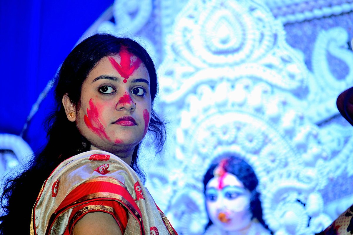 Woman in a cream saree with a red and gold border, her forehead and cheek marked with red sindoor, in front of a white Durga backdrop