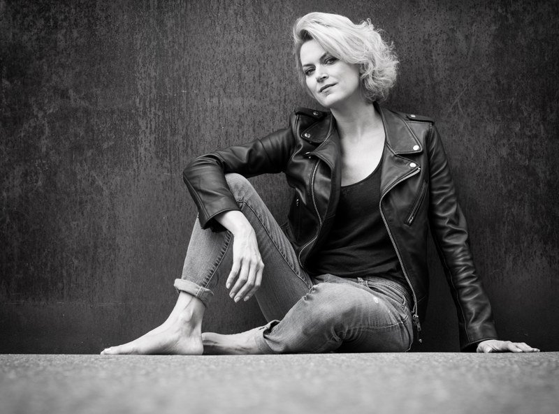 Woman seated in a black leather biker jacket, black top and cuffed jeans.
