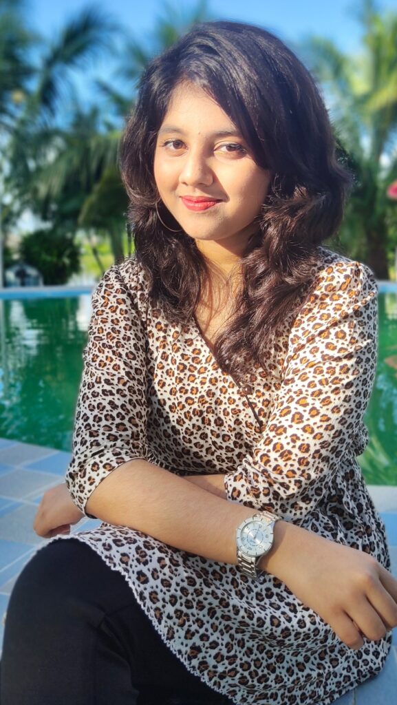 A woman with long dark hair wearing a white leopard-print wrap blouse with three-quarter sleeves and black trousers, seated outdoors by a pool.