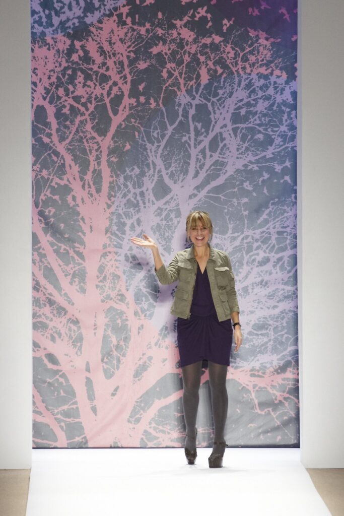 Woman waving on a runway wearing an olive green field jacket over a dark purple wrap dress