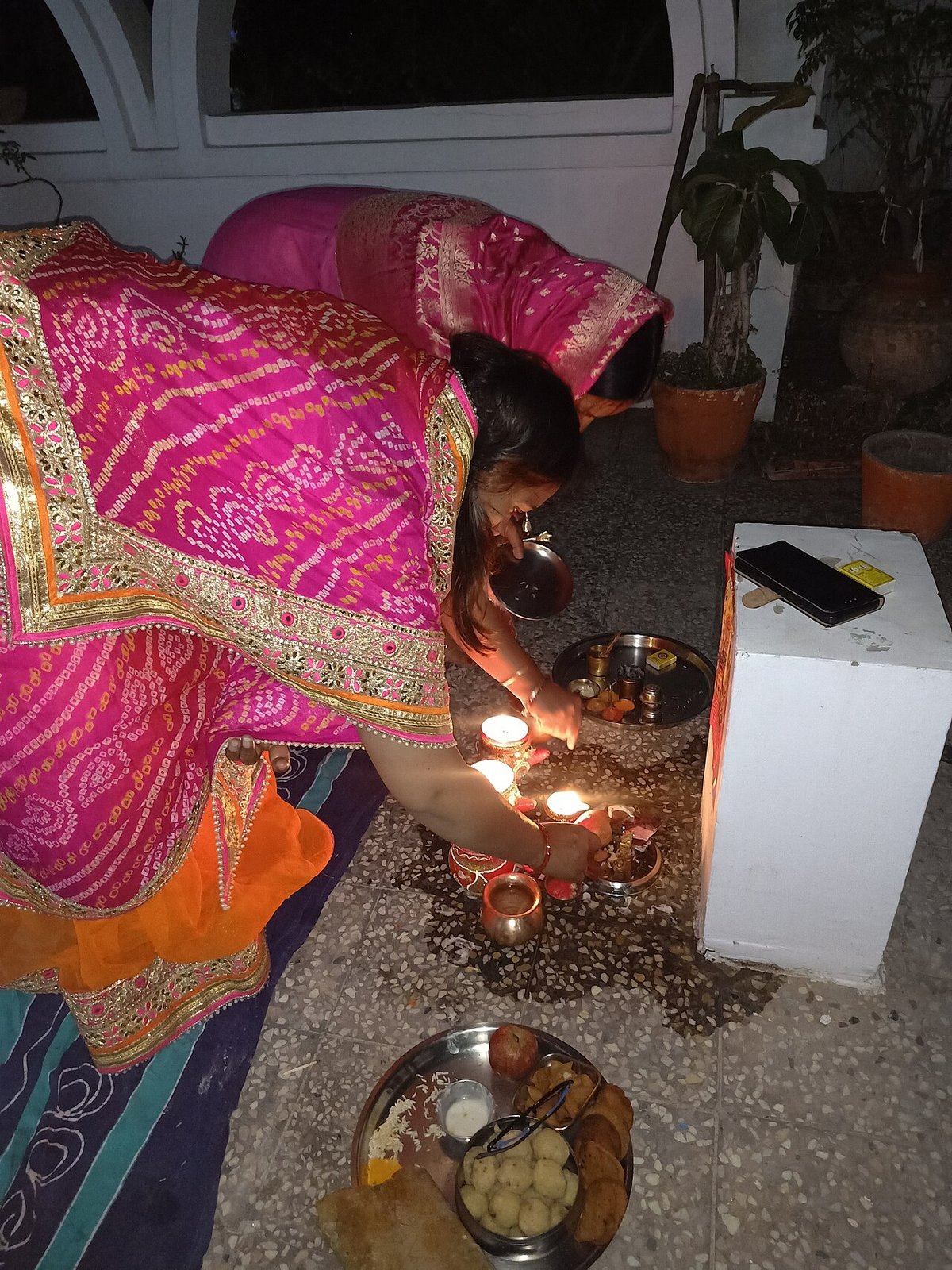Two women in bright pink bandhani sarees with gold borders bending over lit diyas during a Karva Chauth puja at home
