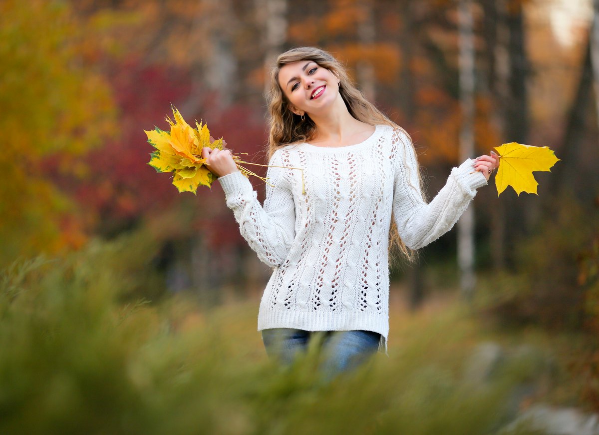Smiling woman outdoors in autumn holding yellow maple leaves, wearing a white openwork knit sweater