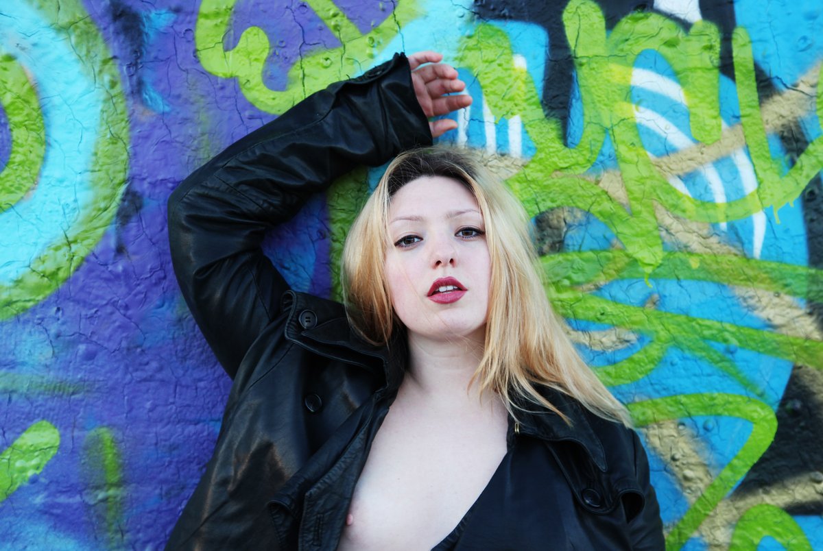 Woman in a black leather jacket standing before a colourful graffiti wall.