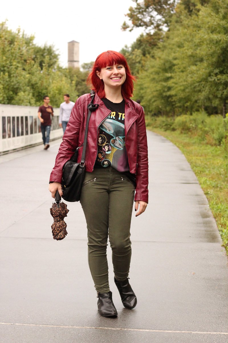 Woman with shoulder-length red hair walking toward the camera on a park path, wearing an open burgundy faux-leather moto jacket over a black graphic tee, olive-green skinny jeans, and black pointed ankle boots, carrying a leopard-print umbrella and a black crossbody bag